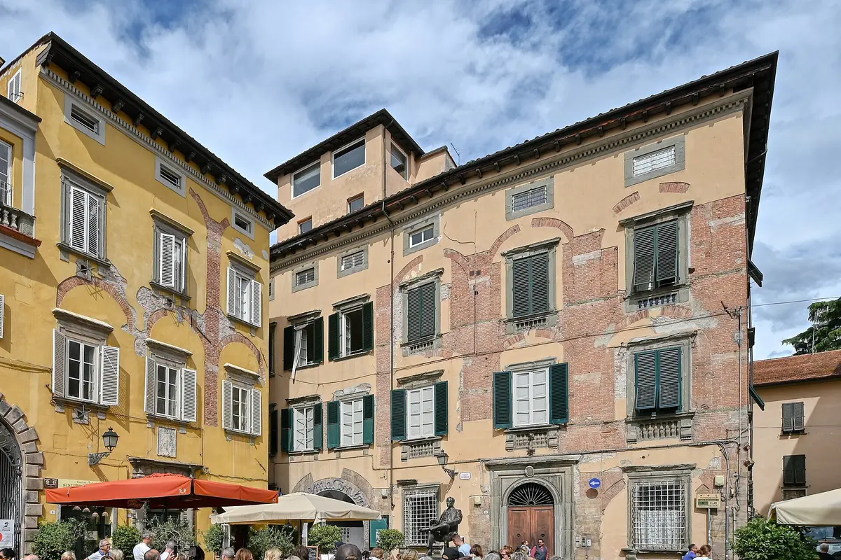 To høje gamle palazzi med okker- og lyserøde pudsvægge og grønne skodder omkring en lille plads i Lucca, med caféparasoller i gadeplan og en blå himmel derover
