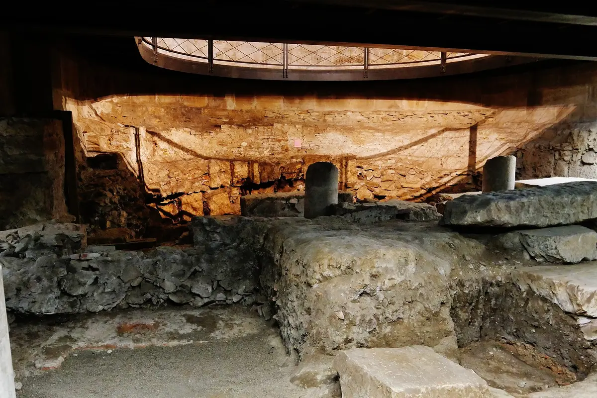A dim excavated chamber beneath a church, with rough walls of Roman and later masonry, two stone column bases and a curved pavement lit by warm lights under the floor above