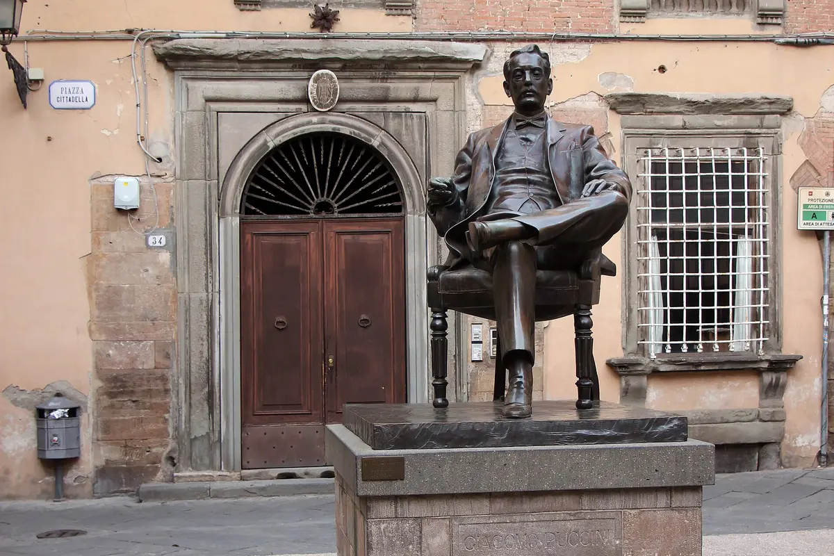 A bronze statue of a seated man in a coat, leaning back in a chair in front of an old arched wooden door on a Lucca street corner.